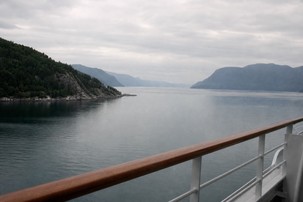 A cruise ship balcony rail with calm fjord water and forested hills beyond, no text, no logos, no people.