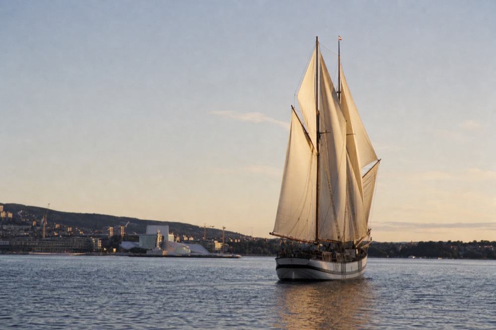 A wooden sailing ship under canvas on the Oslofjord with Oslo's skyline behind it, no text, no logos, no people