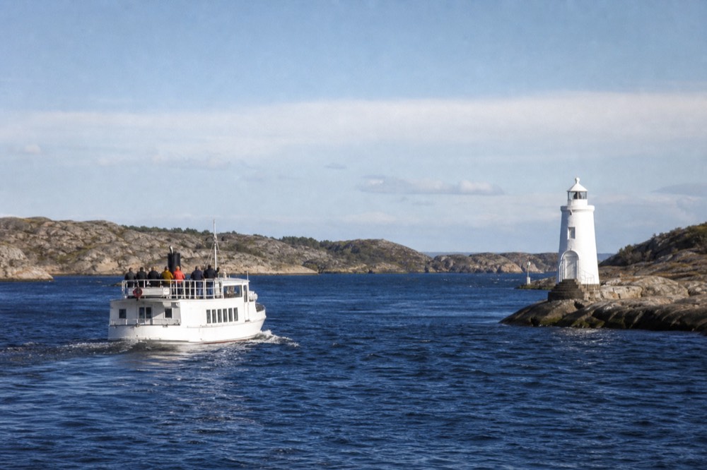 Small white cruise boat passing a lighthouse at the mouth of a narrow fjord channel, no text, no logos, no people