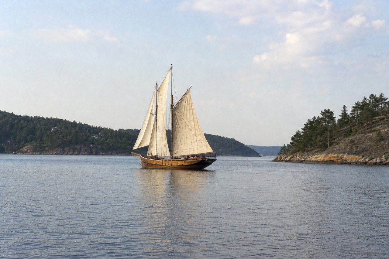 Wooden sailing ship gliding across calm Oslofjord water between green islands, no text, no logos, no people