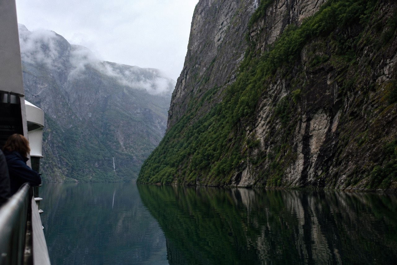 Steep green cliffs dropping into a narrow Norwegian fjord with still water, no text, no logos, no people