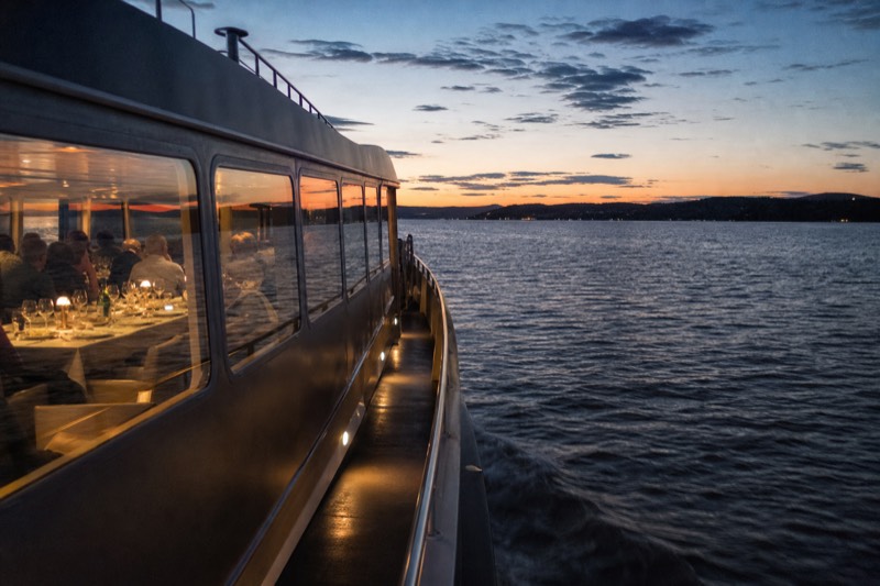 A modern electric-hybrid sightseeing boat crossing the Oslofjord at dusk, a long dinner table set through the wide cabin windows, calm dark water reflecting the sky