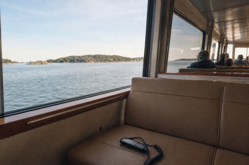 A compact electric sightseeing boat gliding across the Oslofjord near green islands, an audio-guide handset resting on an empty bench seat beside a window