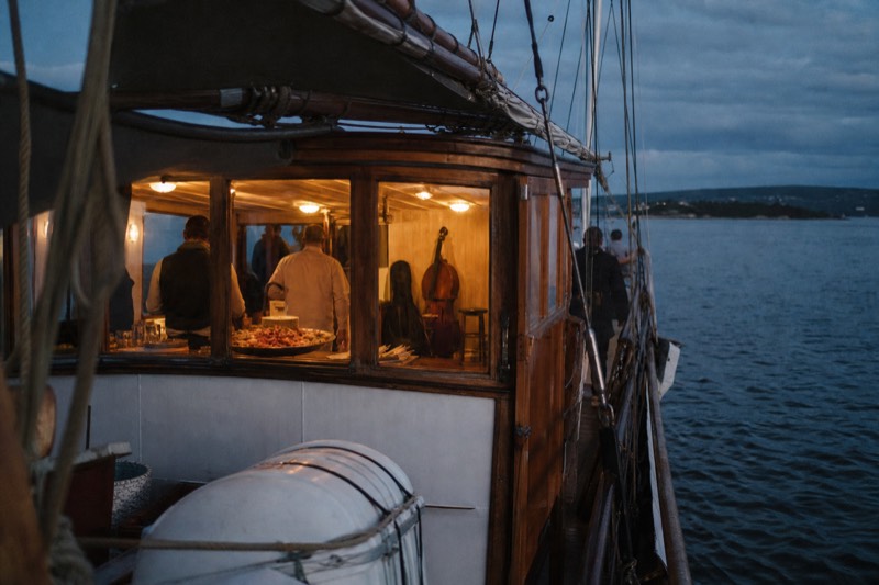 A traditional wooden sailing ship under way on the Oslofjord in the evening, warm light glowing from the cabin where a shrimp buffet and a small musicians' corner are visible
