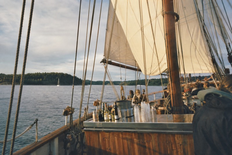 A traditional wooden sailing ship crossing the Oslofjord under full canvas, a small onboard bar counter with drinks visible on deck, green islands in the background