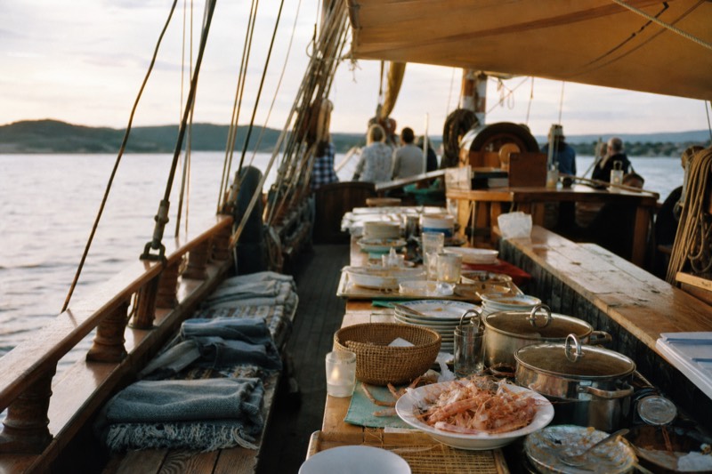 The open deck of a traditional wooden sailing ship on the Oslofjord with a long shrimp buffet table under an awning and folded blankets on the deck benches, golden evening light