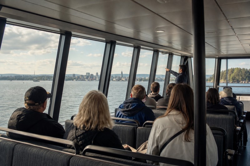 A guide gesturing toward the Oslo skyline from inside a near-silent hybrid-electric sightseeing boat's panoramic cabin, passengers listening from bench seating facing the tall windows
