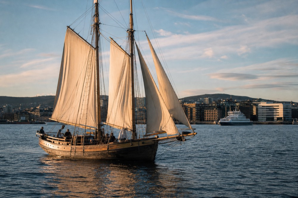 A wooden sailing ship underway on the Oslofjord with Oslo's skyline behind it, no text, no logos, no people